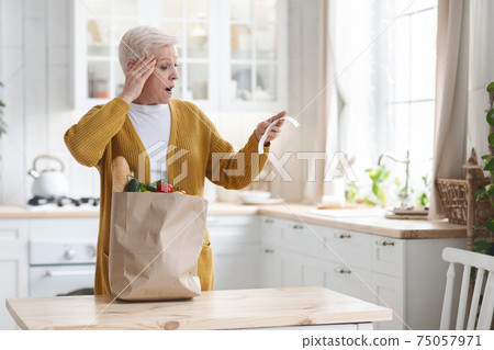 Amazed elderly woman checking grocery bill in kitchen Amazed elderly woman checking grocery bill in kitchen 75057971