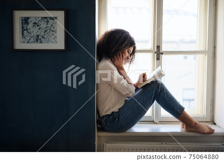 Portrait of young woman with book on window sill indoors at home, reading. 75064880