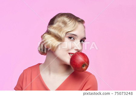 An attractive young blonde with a retro hairstyle holds a red apple in her teeth 75073248