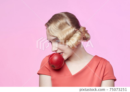 An attractive young blonde with a retro hairstyle holds a red apple in her teeth 75073251