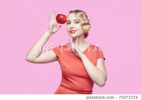 attractive young blonde with a retro hairstyle holds a red apple near her face attractive young blonde with a retro hairstyle holds a red apple near her face 75073259