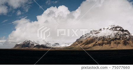 A wide field of dry yellow grass and a mountain in the distance with a snow-capped peak, against a blue sky with white clouds in Iceland. 75077660