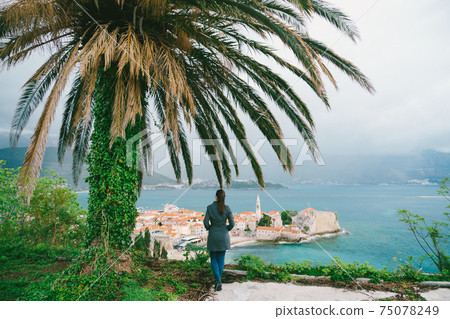 Woman is standing under a palm tree with her back to the camera looking at the Old town of Budva in Montenegro Woman is standing under a palm tree with her back to the camera looking at the Old town of Budva in Montenegro 75078249