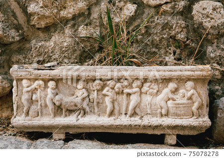 Close-up of a flower pot with a bas-relief depicting people and animals against a stone wall and a green plant. Close-up of a flower pot with a bas-relief depicting people and animals against a stone wall and a green plant. 75078278