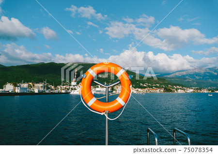 Orange lifebuoy on a metal stand against the backdrop of mountains, water and sky. Orange lifebuoy on a metal stand against the backdrop of mountains, water and sky. 75078354