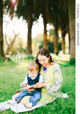 Baby boy is sitting in his mom's lap and reading a book on a picnic in the park Baby boy is sitting in his mom's lap and reading a book on a picnic in the park 75080221