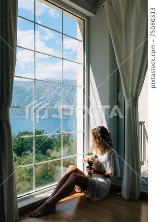 The bride in a white peignoir sits by the window overlooking the Bay of Kotor with a wedding bouquet in her hands  75080313