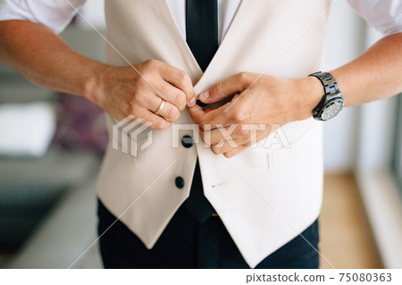A man buttoning the vest of his suit in a hotel room while preparing for a wedding ceremony  75080363