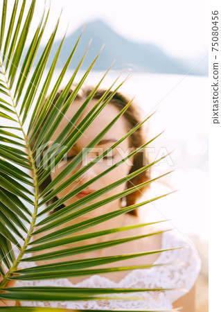 Portrait of a bride with closed eyes through a leaf of a palm tree, behind the sea and a mountain Portrait of a bride with closed eyes through a leaf of a palm tree, behind the sea and a mountain 75080456