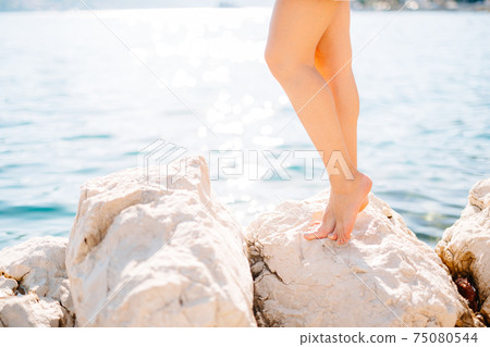 Legs of a woman standing on a stone near the water on a rocky beach Legs of a woman standing on a stone near the water on a rocky beach 75080544
