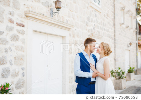 The groom gently hugs the bride against the backdrop of a beautiful white house in Perast  75080558