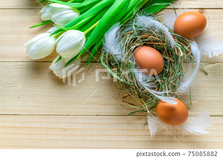 Easter symbol. Natural colour eggs in basket with spring tulips, white feathers on wooden table background in Happy Easter decoration. Congratulatory easter flat lay design. 75085882