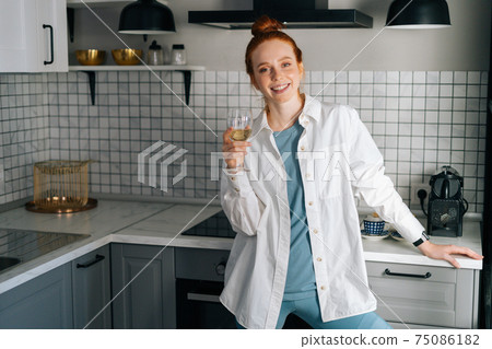 Portrait of cheerful young woman drinking white wine standing in light kitchen room. Portrait of cheerful young woman drinking white wine standing in light kitchen room. 75086182