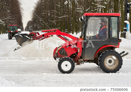 Red tractor with a large bucket removes snow in a city park 75086606