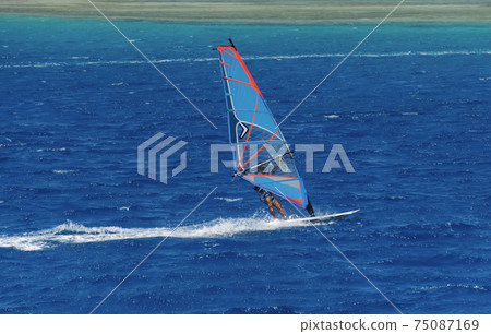the windsurfer on the board under sail moves at a speed along the surface of the sea, against the background of waves and shoreline 75087169