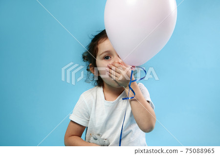 Adorable child girl covers her mouth with her hand and poses to camera behind a pink balloon on blue background with copy space Adorable child girl covers her mouth with her hand and poses to camera behind a pink balloon on blue background with copy space 75088965