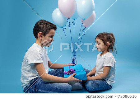 A little boy gives a gift to a little girl sitting in front of him against a background of colored balloons on a blue surface 75090516