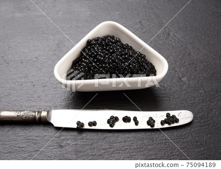 black grainy caviar of paddlefish fish in a white ceramic bowl, black background 75094189