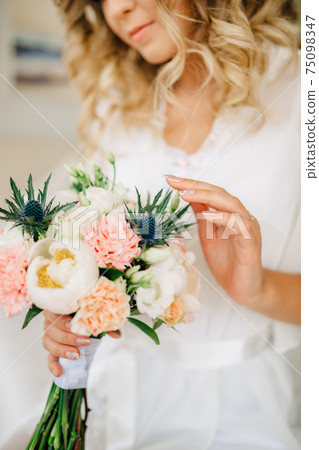 The bride holds in her hand and gently touches a bouquet with peonies, roses, lisianthus and eryngiums  75098347