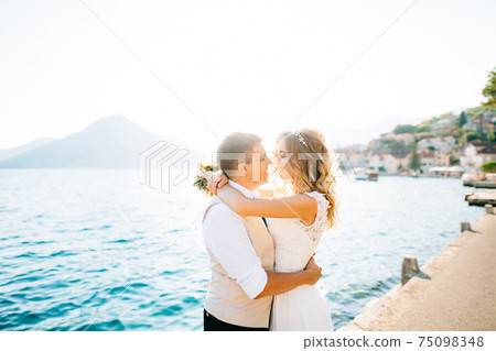 The bride hugs the groom and holds a bouquet in her hands on the pier in the Bay of Kotor  75098348