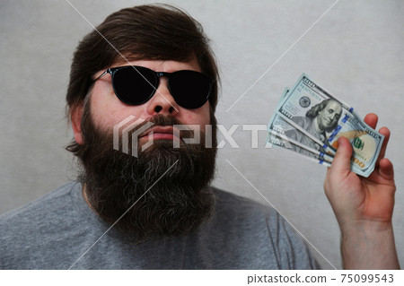 A bearded man with black glasses holds dollars in his hand.  75099543