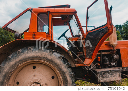 Old red tractor in the field during the haymaking season, pressing hay on bales, forage harvesting. 75101042