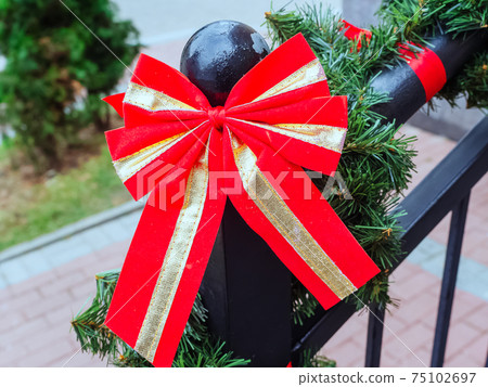 A red-gold bow hangs on a metal railing wrapped in an artificial spruce branch. Close-up photo A red-gold bow hangs on a metal railing wrapped in an artificial spruce branch. Close-up photo 75102697