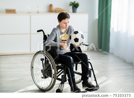 Teen handicapped soccer player with ball and trophy sitting in wheelchair, feeling depressed over his injury at home 75103373