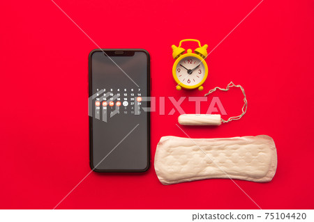 Top view of menstruation calendar in smartphone with cotton tampon, sanitary pad and yellow alarm clock. Woman critical days, woman hygiene protection 75104420
