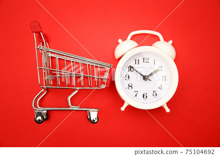 Mini shopping cart with white alarm clock on red background. Conceptual image of sale, seasonal discounts in shopping stores, discount time, Time saving concept 75104692