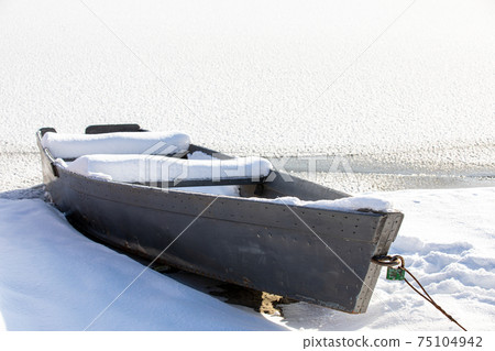 Snow-covered old wooden rowing boat on the shore in winter season. Fishing boat. A frozen lake or river. Low ambient temperature. The season of winter fishing. Sunny winter day. 75104942