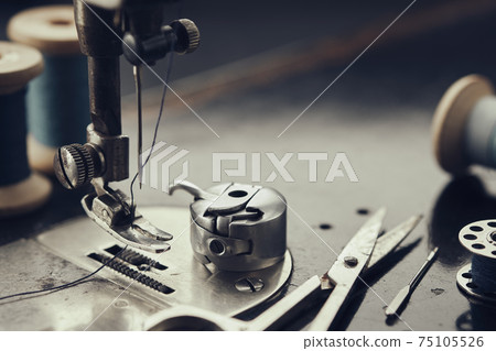 Close up on metal shuttle, foot of vintage sewing machine, scissors, spool of thread, sewing needles. Sewing items on working part of antique sewing machine. Close up on metal shuttle, foot of vintage sewing machine, scissors, spool of thread, sewing needles. Sewing items on working part of antique sewing machine. 75105526