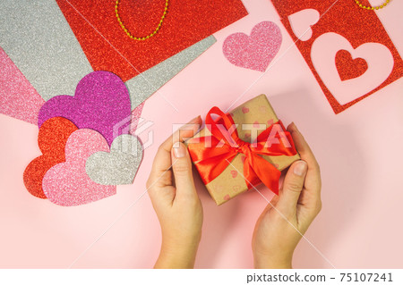 Packaging Valentine's Day or birthday presents. Valentine's Day gifts with a red paper heart on a pink background. Top view. Female hands hold gift box with red ribbon. Packaging Valentine's Day or birthday presents. Valentine's Day gifts with a red paper heart on a pink background. Top view. Female hands hold gift box with red ribbon. 75107241