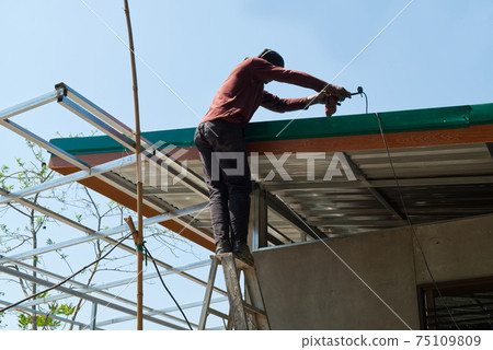 Unsafe Male Roofer Workman Using Electric Screwdriver Install Tile on Roof of New House in the Construction Site with no Protection 75109809