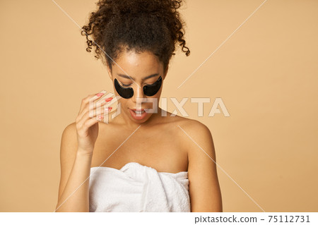 Portrait of mixed race young woman after shower posing with applied black beauty patches under eyes isolated over beige background Portrait of mixed race young woman after shower posing with applied black beauty patches under eyes isolated over beige background 75112731