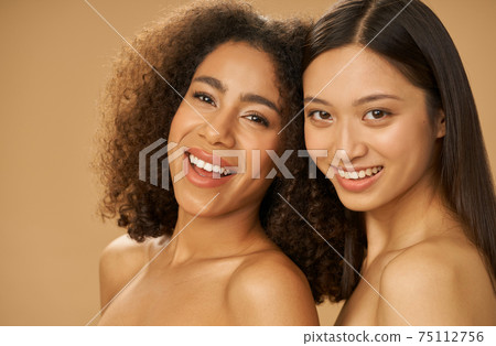 Portrait of two joyful attractive mixed race young women with perfect smile posing for camera isolated over beige background 75112756