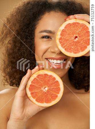 Portrait of mixed race young woman looking happy while posing with grapefruit cut in half over beige background 75112759