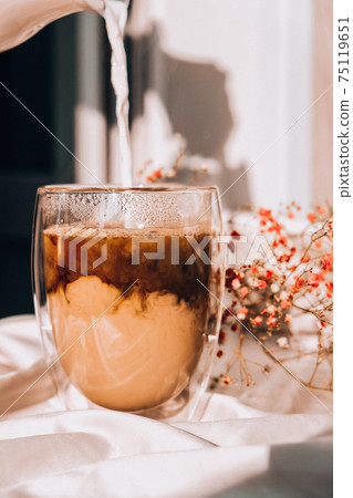 Milk pouring into the glass cup with black coffee. Morning aesthetics vibes. Breakfast. Pink gypsophila flowers 75119651