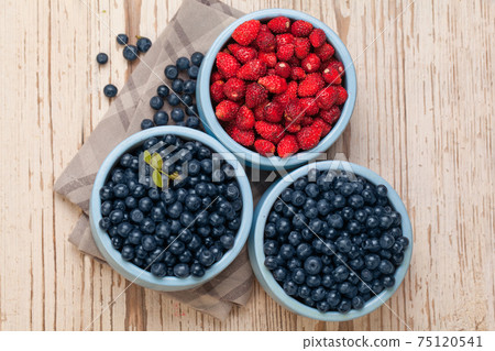 Bilberry and strawberry in blue bowls on white vintage wooden background, top view food composition 75120541