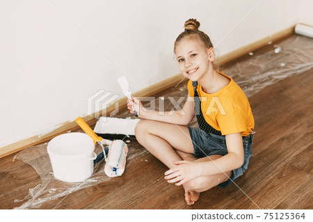 A girl in a denim overalls and a yellow T-shirt helps to paint the walls in an apartment white A girl in a denim overalls and a yellow T-shirt helps to paint the walls in an apartment white 75125364