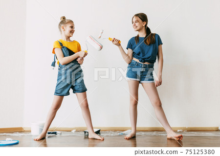 A girl with her sister in bright blue and yellow clothes helps to paint the walls in her room white A girl with her sister in bright blue and yellow clothes helps to paint the walls in her room white 75125380