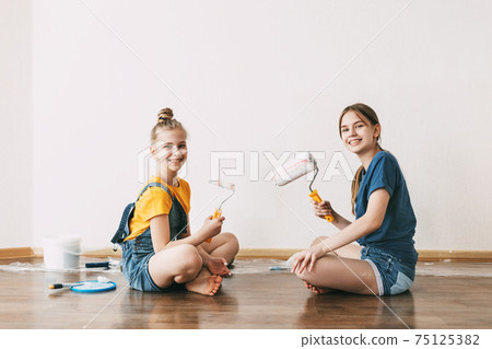 A girl with her sister in bright blue and yellow clothes helps to paint the walls in her room white A girl with her sister in bright blue and yellow clothes helps to paint the walls in her room white 75125382
