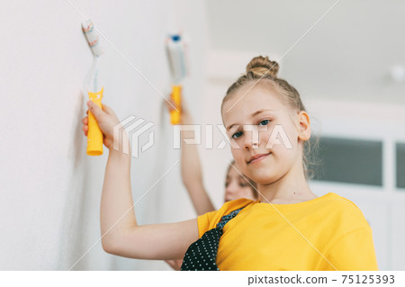 A girl with her sister in bright blue and yellow clothes helps to paint the walls in her room white 75125393