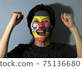 Cheerful portrait of a man with the flag of Uganda painted on his face on grey background. 75136680