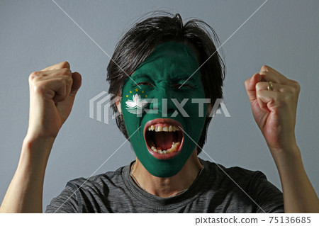Cheerful portrait of a man with the flag of the Macau painted on his face on grey background. 75136685