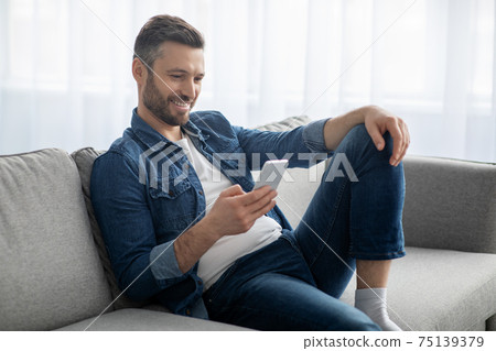 Happy man sitting on couch at home, using mobile phone 75139379