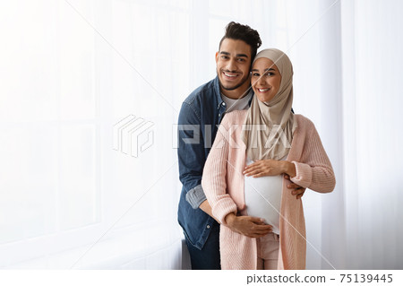 Pregnant Muslim Woman And Her Husband Embracing Near Window At Home Pregnant Muslim Woman And Her Husband Embracing Near Window At Home 75139445