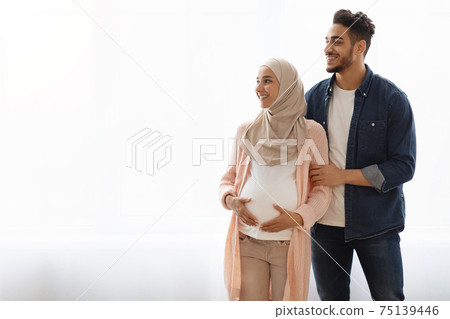 Pregnant Religious Muslim Couple Standing Near Window At Home, Looking Aside Pregnant Religious Muslim Couple Standing Near Window At Home, Looking Aside 75139446