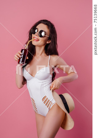 tanned young woman in straw hat, white swimsuit hold a cocktail on pink background, isolate 75139668