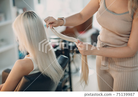 Hairdresser female making hair extensions to young woman with blonde hair in beauty salon. Professional hair extension. Hairdresser female making hair extensions to young woman with blonde hair in beauty salon. Professional hair extension. 75139987